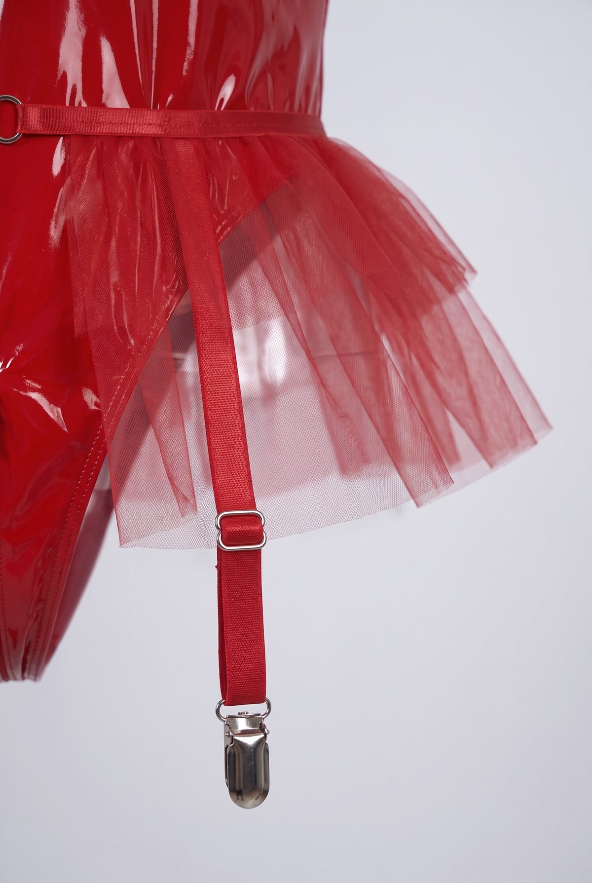 Close-up of the glossy vinyl waist, tulle peplum and garter strap clip of the midnight mesh temptation bodysuit in red